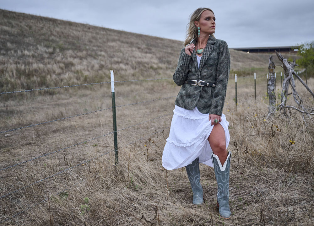 A woman standing in a pasture with tall grass, posing in a chic jacket, looking away from the camera.